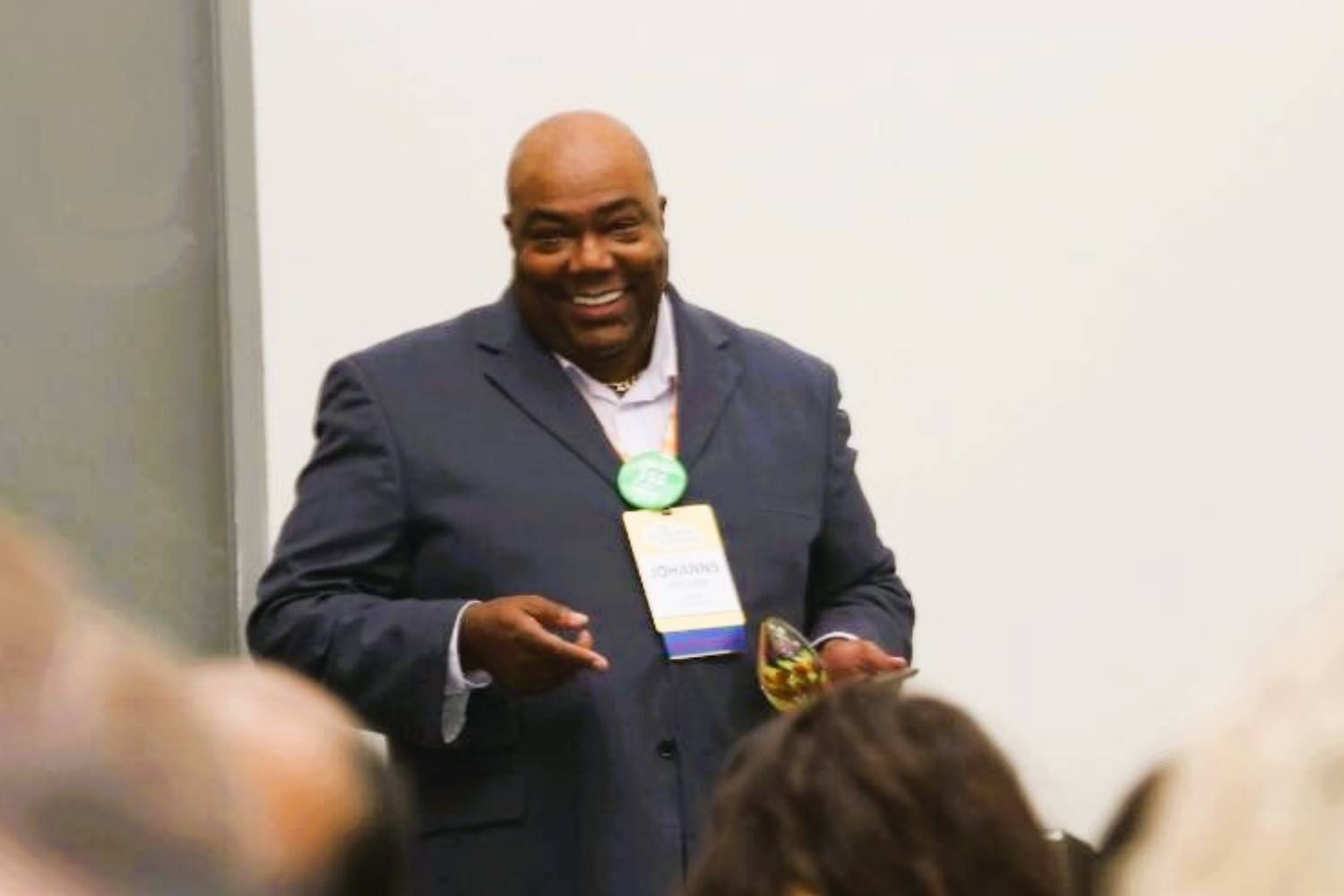 A man in a suit stands in front of an audience, holding an object and gesturing, with a name tag visible on his jacket.